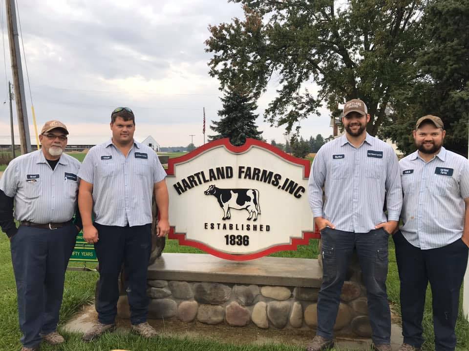 Four employees stand proudly in front of the Hartland Farms, Inc. sign, established in 1836, showcasing the farm's long history. The group is dressed in matching uniforms, emphasizing their commitment to the agricultural industry. The setting features a well-maintained lawn and trees, reflecting the farm's dedication to quality and tradition.