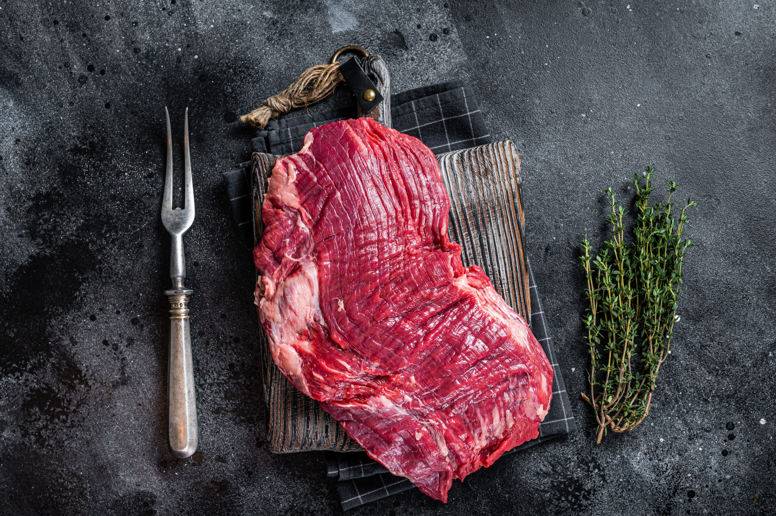 Fresh raw flank steak displayed on a wooden cutting board with a silver meat fork and sprigs of thyme, set against a dark textured background. Ideal for culinary recipes and meat preparation tips.