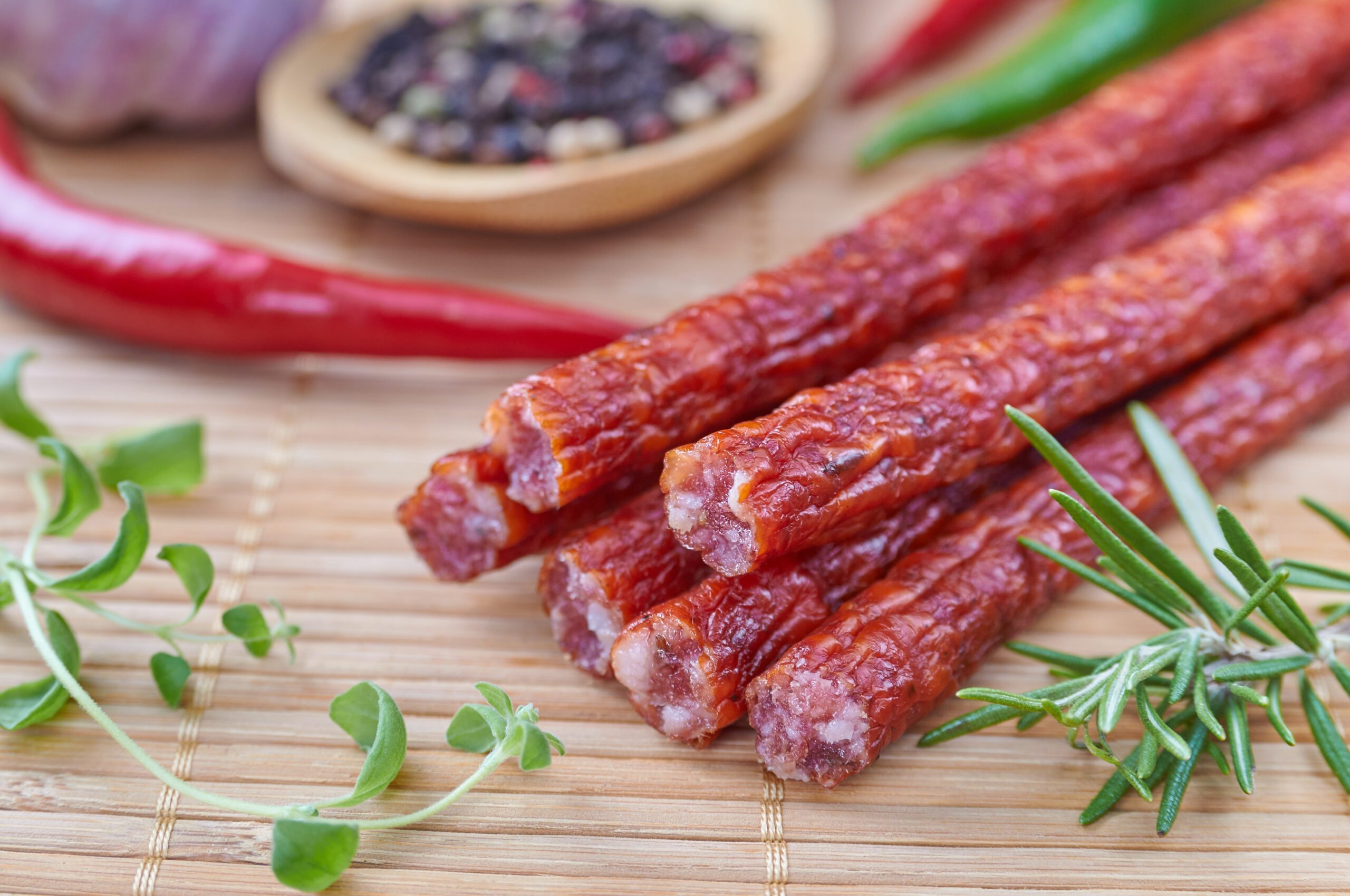 Close-up of dried meat sticks arranged on a bamboo mat, surrounded by fresh herbs like oregano and rosemary, with red and green chili peppers and a bowl of mixed peppercorns in the background, highlighting the ingredients for a flavorful snack.