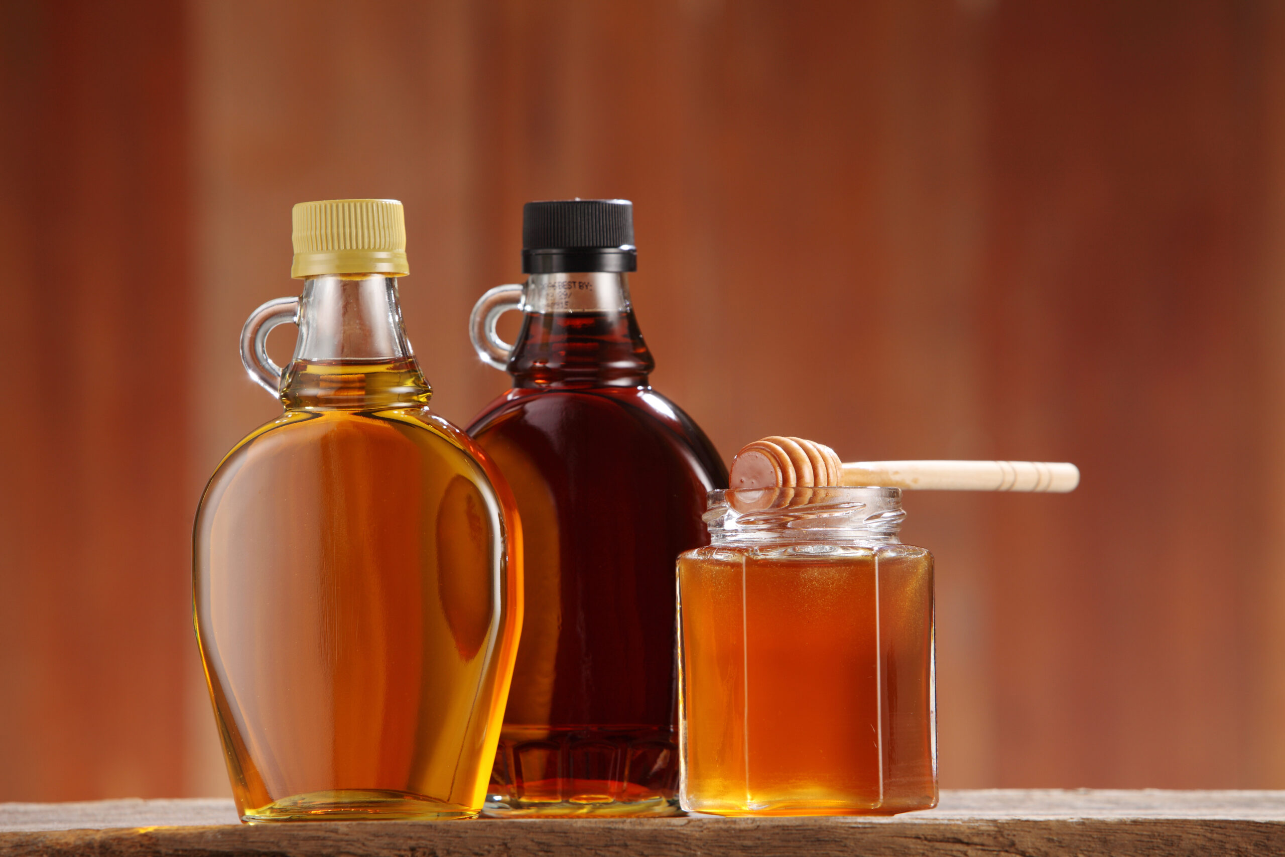 Bottles of maple syrup and a jar of honey with a wooden dipper on a wooden surface, showcasing natural sweeteners.